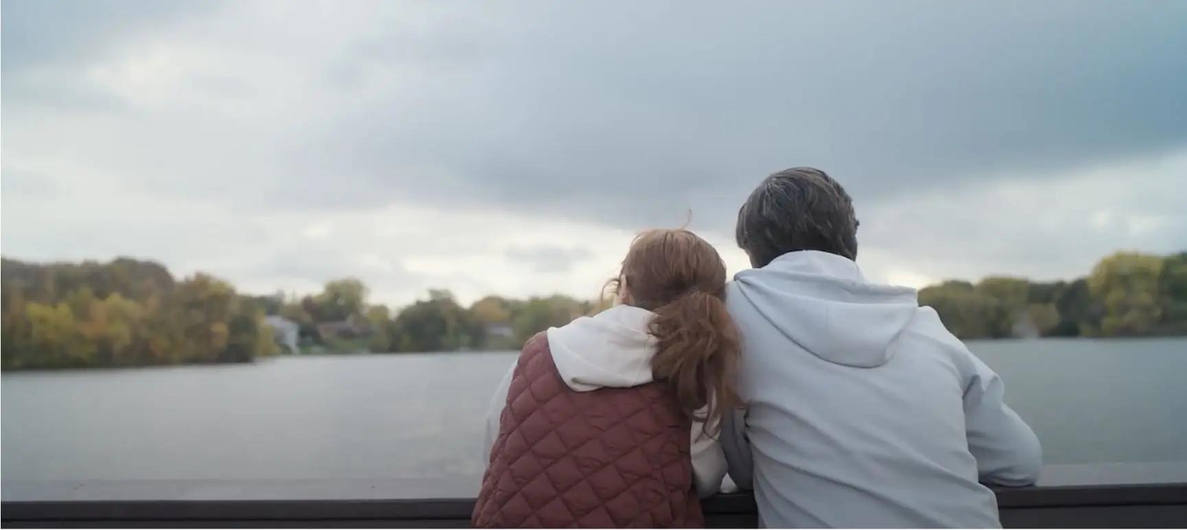 The image shows two individuals sitting closely together, looking out over a body of water.