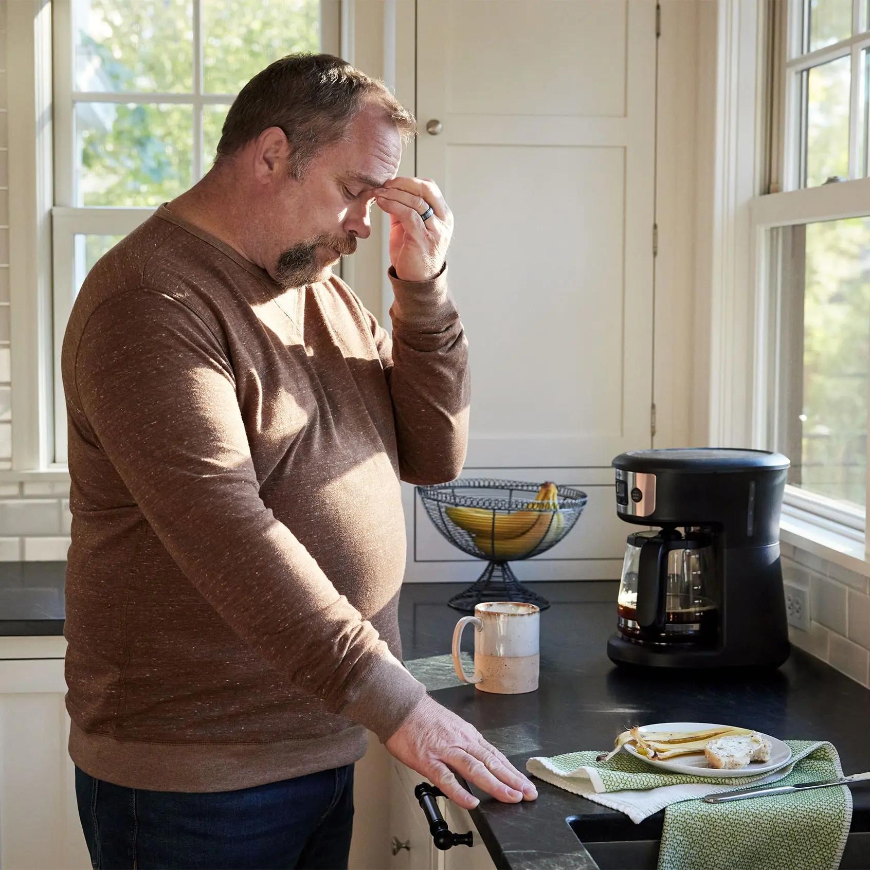 The image shows a man standing in a kitchen, holding his forehead indicating fatigue.